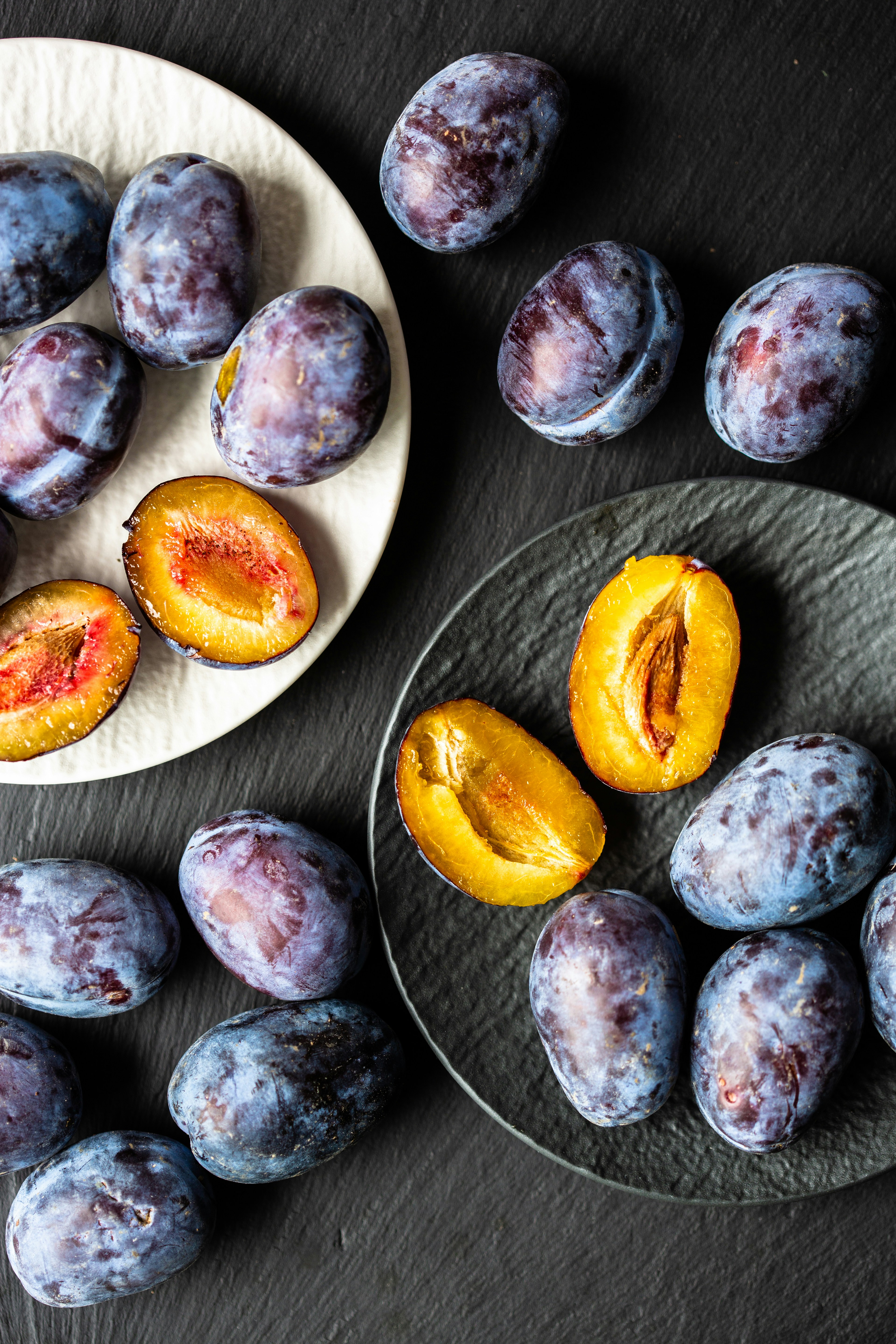 Ripe fresh organic plums on a dark kitchen counter on two textured plates. Blue and purple on black and white.