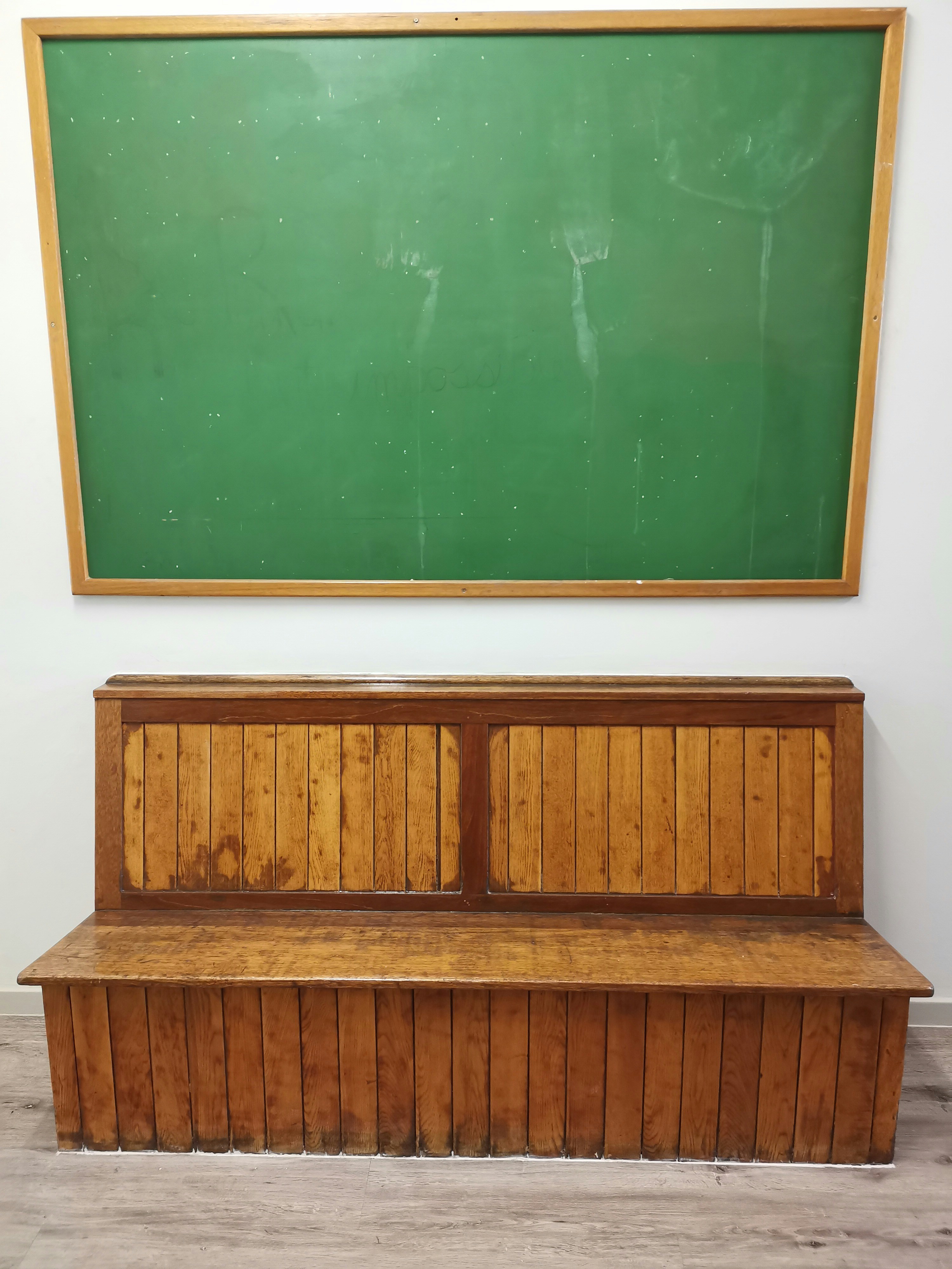Green chalkboard mounted above a weathered wooden bench in a classroom. The scene emphasizes aged wood textures and subdued colors.
