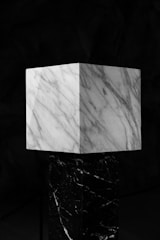 A marble cube with distinct veins sits atop a pedestal of darker marble against a dark background. The cube has a light gray and white pattern, creating a striking contrast with the black and white-veined pedestal.