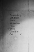 A concrete wall with a list of locations and events written in both German and English. The text includes 'Ausstellung', 'Exhibition', 'Event', 'Workshop', 'Foyer', 'Shop', 'Café/Bar', and 'Exit'. An arrow points to the right, indicating direction.