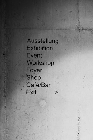 A concrete wall with a list of locations and events written in both German and English. The text includes 'Ausstellung', 'Exhibition', 'Event', 'Workshop', 'Foyer', 'Shop', 'Café/Bar', and 'Exit'. An arrow points to the right, indicating direction.