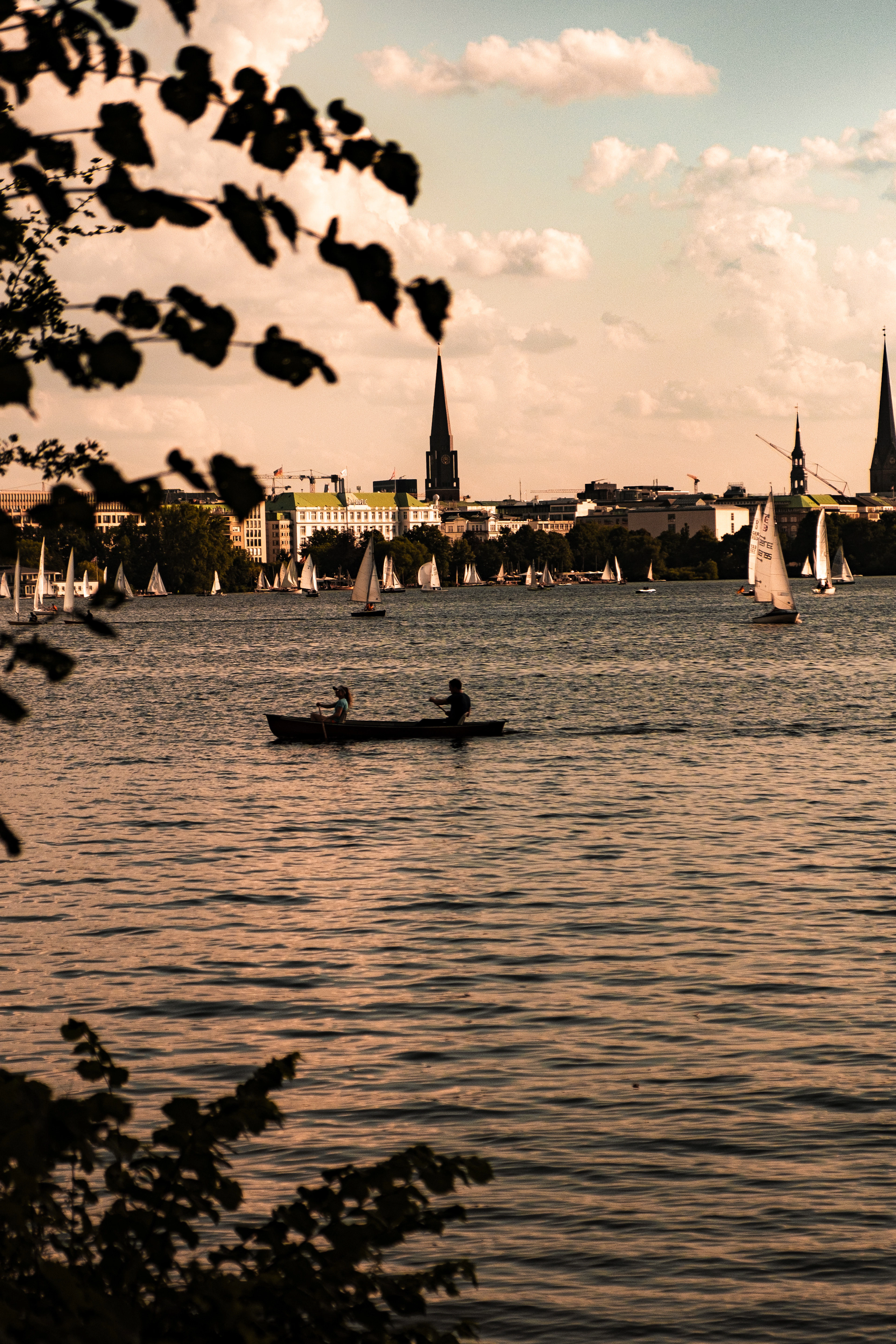 Two people rowing a small wooden boat on Hamburg's Aussenalster in late afternoon light, a fleet of white dinghy sails behind them and the city's church spires along the far shore, framed by overhanging leaves