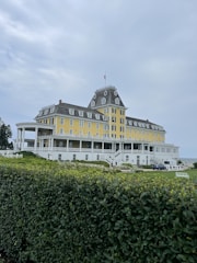 A large, historic hotel with a yellow facade and grey roof sits atop a hill. The building has multiple windows and a porch that wraps around the front. A manicured green hedge is in the foreground, with a cloudy sky overhead. There are a few people and some vehicles visible around the premises.