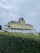 A large, historic hotel with a yellow facade and grey roof sits atop a hill. The building has multiple windows and a porch that wraps around the front. A manicured green hedge is in the foreground, with a cloudy sky overhead. There are a few people and some vehicles visible around the premises.