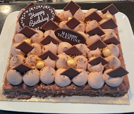A rectangular cake decorated with chocolate pieces, dollops of frosting, and gold-colored spheres. The cake has two chocolate plaques, one with the words 'Happy Birthday' and the other with 'Maison Valentine'. The cake sits on a white plate.