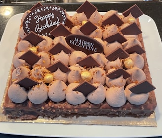 A rectangular cake decorated with chocolate pieces, dollops of frosting, and gold-colored spheres. The cake has two chocolate plaques, one with the words 'Happy Birthday' and the other with 'Maison Valentine'. The cake sits on a white plate.