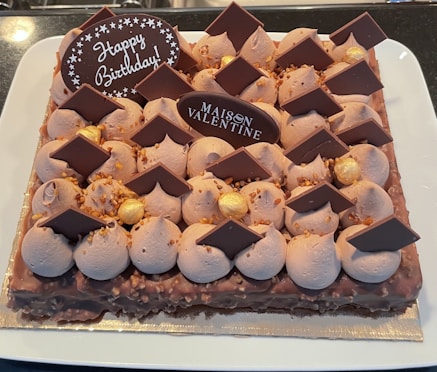 A rectangular cake decorated with chocolate pieces, dollops of frosting, and gold-colored spheres. The cake has two chocolate plaques, one with the words 'Happy Birthday' and the other with 'Maison Valentine'. The cake sits on a white plate.