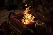 A brick altar adorned with colorful garlands of pink and yellow flowers surrounds a vibrant, dancing flame. Several people are gathered around, participating in what appears to be a traditional ritual. In front of the altar, a bowl with a shiny liquid is placed, possibly for ceremonial use. Some items such as lamps and offerings are visible, adding to the spiritual atmosphere.