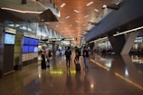 A modern airport terminal with high ceilings and long corridors. Information displays and signboards direct travelers towards gates C1-C29. People are moving with luggage, some checking flight information on the screens.