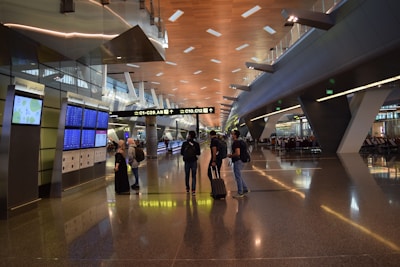 A modern airport terminal with high ceilings and long corridors. Information displays and signboards direct travelers towards gates C1-C29. People are moving with luggage, some checking flight information on the screens.