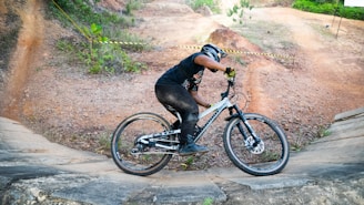 A person wearing a helmet is skillfully riding a mountain bike on a dirt trail with curved slopes. The surroundings include patches of greenery and orange-brown soil. The cyclist is dressed in black with gloves and is positioned in action as if navigating a turn.