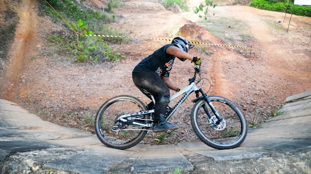 A person wearing a helmet is skillfully riding a mountain bike on a dirt trail with curved slopes. The surroundings include patches of greenery and orange-brown soil. The cyclist is dressed in black with gloves and is positioned in action as if navigating a turn.