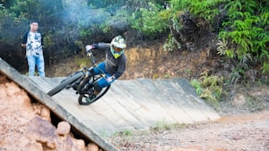A mountain biker in full gear is maneuvering sharply on a dirt track ramp while another person stands observing nearby. The lush greenery and uneven terrain emphasize an outdoor adventure setting.