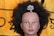 A mannequin head with dark, curly hair adorned with a colorful, jeweled hairpiece shaped like a tree. The backdrop is a fabric with a yellow and black pattern.