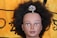 A mannequin head with dark, curly hair adorned with a colorful, jeweled hairpiece shaped like a tree. The backdrop is a fabric with a yellow and black pattern.