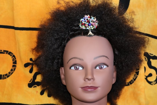 A mannequin head with dark, curly hair adorned with a colorful, jeweled hairpiece shaped like a tree. The backdrop is a fabric with a yellow and black pattern.