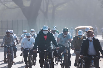 A group of people wearing masks and helmets are riding bicycles on a hazy day. They appear to be participating in an organized event or cycling activity. The background shows trees and some urban elements like a vehicle.