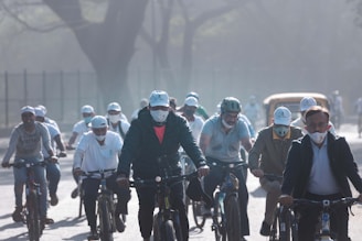 A group of people wearing masks and helmets are riding bicycles on a hazy day. They appear to be participating in an organized event or cycling activity. The background shows trees and some urban elements like a vehicle.