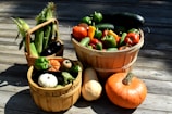 Several baskets of fresh vegetables are placed on a wooden deck. The largest basket is filled with a variety of colorful peppers and cucumbers. Another basket contains corn with husks and shiny eggplants, while a smaller basket labeled 'Welcome' holds a selection of small pumpkins and squashes. An orange pumpkin and a butternut squash are positioned next to the baskets.