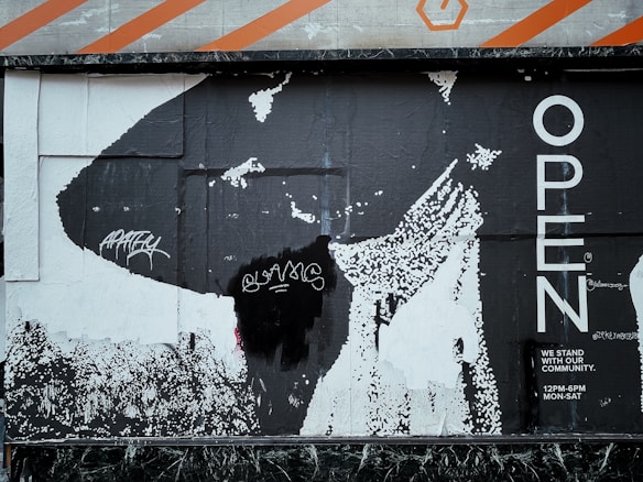 A black and white poster with bold lettering saying 'OPEN' on the right. The background includes abstract graphics with a large black shape and some pixelated areas. There are graffiti tags in white on the left side, and the text 'WE STAND WITH OUR COMMUNITY. 12PM-6PM MON-SAT' is visible in the bottom right corner.