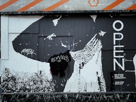 A black and white poster with bold lettering saying 'OPEN' on the right. The background includes abstract graphics with a large black shape and some pixelated areas. There are graffiti tags in white on the left side, and the text 'WE STAND WITH OUR COMMUNITY. 12PM-6PM MON-SAT' is visible in the bottom right corner.