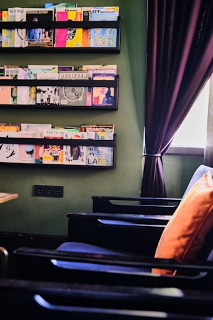 A cozy reading nook featuring black shelves on a green wall, filled with an assortment of magazines. Below the shelves, a row of dark wooden chairs with an orange cushion add warmth to the space. A purple curtain partially covers a window, creating a relaxed atmosphere.