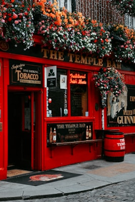 A vibrant red pub facade adorned with an array of colorful hanging flowers above the entrance. The signage reads 'The Temple Bar' with advertisements for tobacco and traditional Irish music. A red barrel branded with the bar's name is near the entrance, and cobblestone pavement is visible in front.