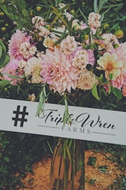 A lush bouquet of various flowers, including pink and cream-colored dahlias, is arranged in a tall clear vase. The bouquet is placed outdoors on a natural ground setting, amid surrounding greenery. A sign reading 'Triple Wren Farms' is placed in front of the arrangement, highlighting a hashtag symbol.