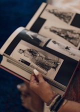 A person is holding open a book featuring black and white photographs of fashion designs. The person’s hand is visible with white nail polish and wrist accessories. The book is resting on a surface, and the background includes a textured fabric.