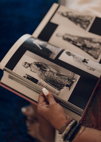 A person is holding open a book featuring black and white photographs of fashion designs. The person&rsquo;s hand is visible with white nail polish and wrist accessories. The book is resting on a surface, and the background includes a textured fabric.