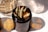 A glass jar holds several rolled items resembling small paper tubes. The jar has a dark amber hue, and sunlight creates intricate patterns on a white surface beside it. Another similar object is partially visible in the background, reflecting the light.