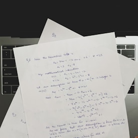 Several sheets of paper with handwritten mathematical notes are placed on a laptop keyboard. The notes include equations and text related to recurrence relations and mathematical induction, written in blue ink. The layout suggests a focus on problem-solving or academic work.