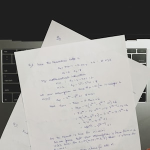 Several sheets of paper with handwritten mathematical notes are placed on a laptop keyboard. The notes include equations and text related to recurrence relations and mathematical induction, written in blue ink. The layout suggests a focus on problem-solving or academic work.