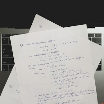 Several sheets of paper with handwritten mathematical notes are placed on a laptop keyboard. The notes include equations and text related to recurrence relations and mathematical induction, written in blue ink. The layout suggests a focus on problem-solving or academic work.