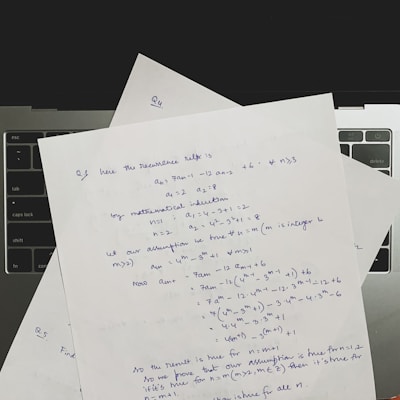 Several sheets of paper with handwritten mathematical notes are placed on a laptop keyboard. The notes include equations and text related to recurrence relations and mathematical induction, written in blue ink. The layout suggests a focus on problem-solving or academic work.