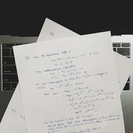 Several sheets of paper with handwritten mathematical notes are placed on a laptop keyboard. The notes include equations and text related to recurrence relations and mathematical induction, written in blue ink. The layout suggests a focus on problem-solving or academic work.