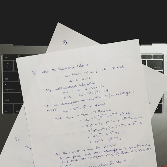 Several sheets of paper with handwritten mathematical notes are placed on a laptop keyboard. The notes include equations and text related to recurrence relations and mathematical induction, written in blue ink. The layout suggests a focus on problem-solving or academic work.