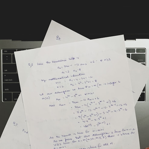 Several sheets of paper with handwritten mathematical notes are placed on a laptop keyboard. The notes include equations and text related to recurrence relations and mathematical induction, written in blue ink. The layout suggests a focus on problem-solving or academic work.