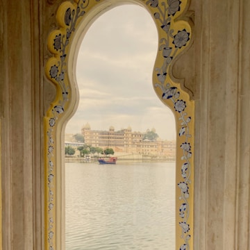 A scenic view through an ornate archway adorned with floral designs, looking onto a serene body of water with a large historic palace or fort in the background. A small boat can be seen on the water, adding to the picturesque quality of the scene.