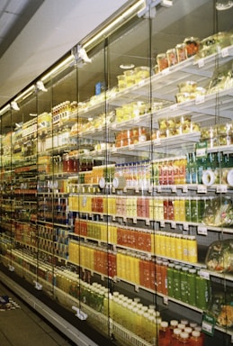 A store aisle featuring a large refrigerated section stocked with a colorful assortment of bottled beverages and packaged fruit cups. The products are neatly arranged on shelves behind glass doors, illuminated by bright indoor lighting.