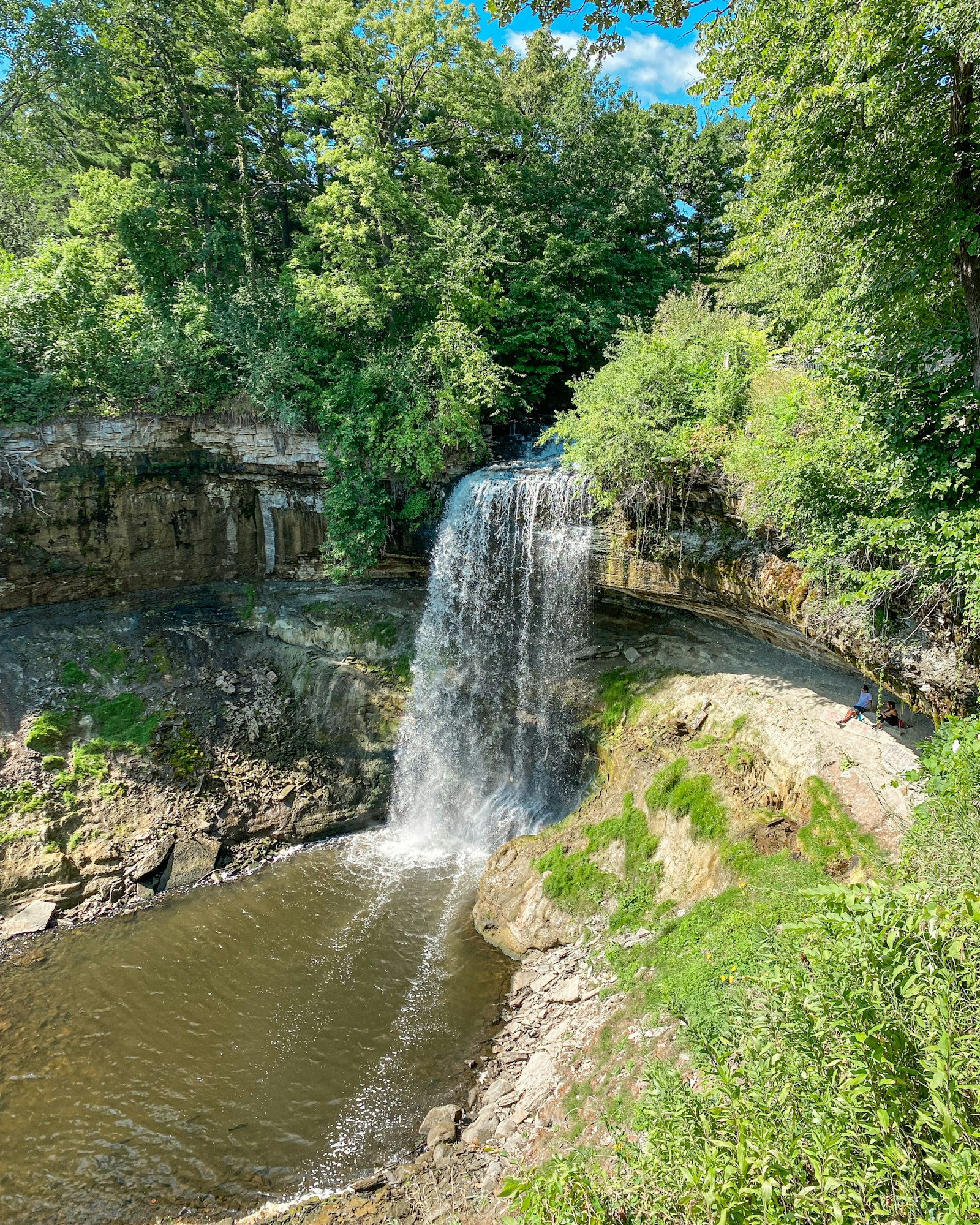 Minnehaha Falls pouring over a limestone ledge into a rocky pool ringed by dense green summer foliage