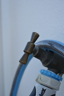 A close-up view of a garden hose attached to a faucet with a brass valve. The hose is blue with a mesh-like texture and connects to a nozzle labeled with text partially visible at the bottom. The background is a plain white surface.