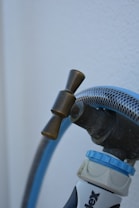 A close-up view of a garden hose attached to a faucet with a brass valve. The hose is blue with a mesh-like texture and connects to a nozzle labeled with text partially visible at the bottom. The background is a plain white surface.