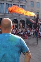 A fire performer on a unicycle breathes a large flame in front of a crowd. The scene is outdoors with an audience gathered in front of an old building. A bald man wearing a blue tie-dye shirt and a tribal armband tattoo stands in the foreground, observing the performance.