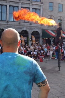 A fire performer on a unicycle breathes a large flame in front of a crowd. The scene is outdoors with an audience gathered in front of an old building. A bald man wearing a blue tie-dye shirt and a tribal armband tattoo stands in the foreground, observing the performance.