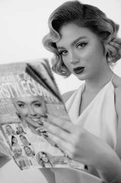 A person with vintage-style makeup and hair is holding a fashion magazine. The person has bold lipstick and eyeliner, and there is a focus on their glamorous appearance. The magazine has multiple celebrity images on the cover.