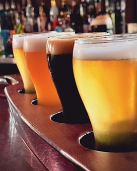 Four distinct glasses of beer with varying colors are lined up in a wooden tray, with a blurred backdrop of bottles on shelves, creating a cozy and inviting bar atmosphere.