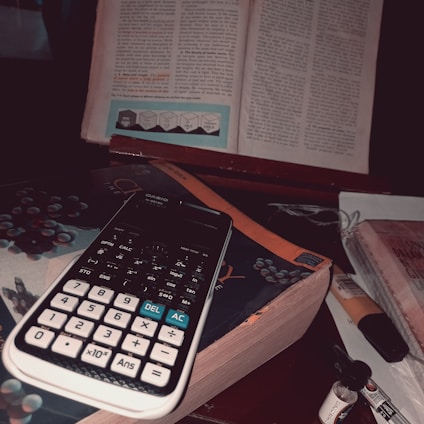 A calculator is placed on top of a stack of books, one of which is titled 'Chemistry'. An open book with text and diagrams is positioned in the background, suggesting a study or work environment. There are additional items like a small bottle and a tube nearby.