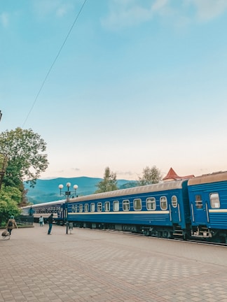 A blue train is stationed on a platform with a scenic backdrop of mountains and trees. Several people are walking along the platform, and a lamppost with spherical lights is visible. The sky is light blue with some scattered clouds.