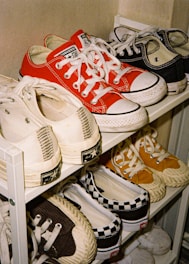 A collection of various sneakers is neatly displayed on a white rack with multiple shelves. The footwear includes classic Converse All Star sneakers in red, black, white, and off-white colors, along with a pair of checkered Vans. Each pair is arranged side by side, showcasing their different designs and colors.